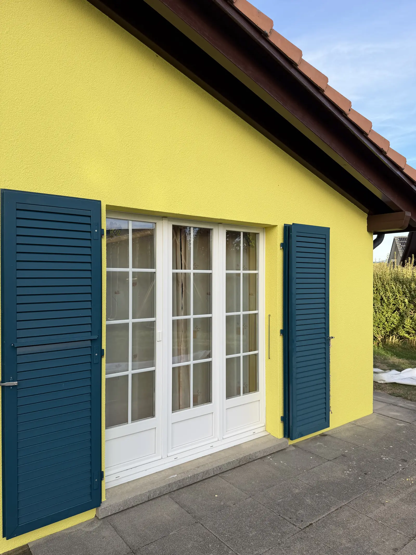 Crépi repeint en jaune citron au ras des cadres blancs de la porte-fenêtre, entre les deux volets